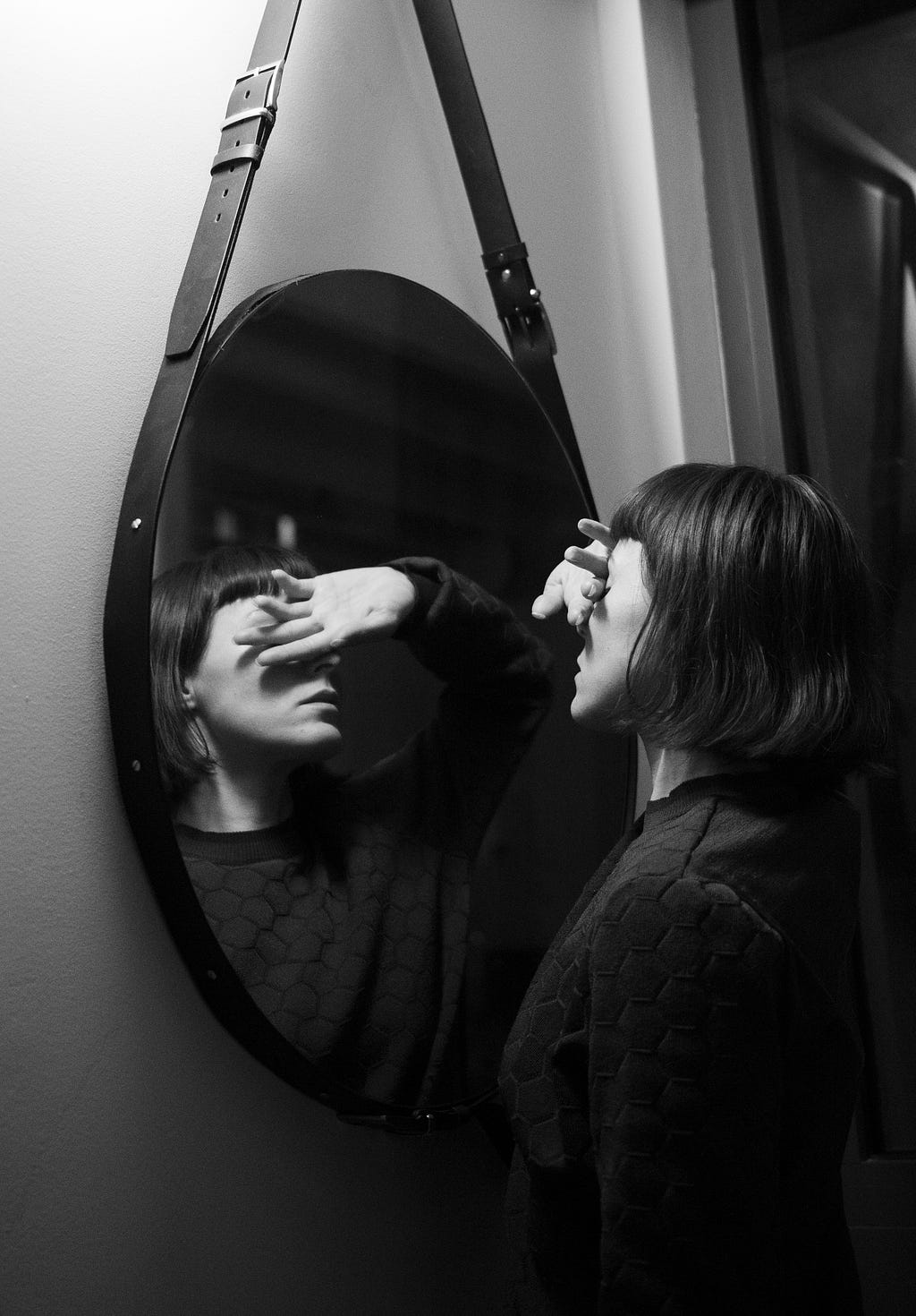A black & white image of a lady looking into a round hanging mirror covering her eyes to stop her from seeing her reflection