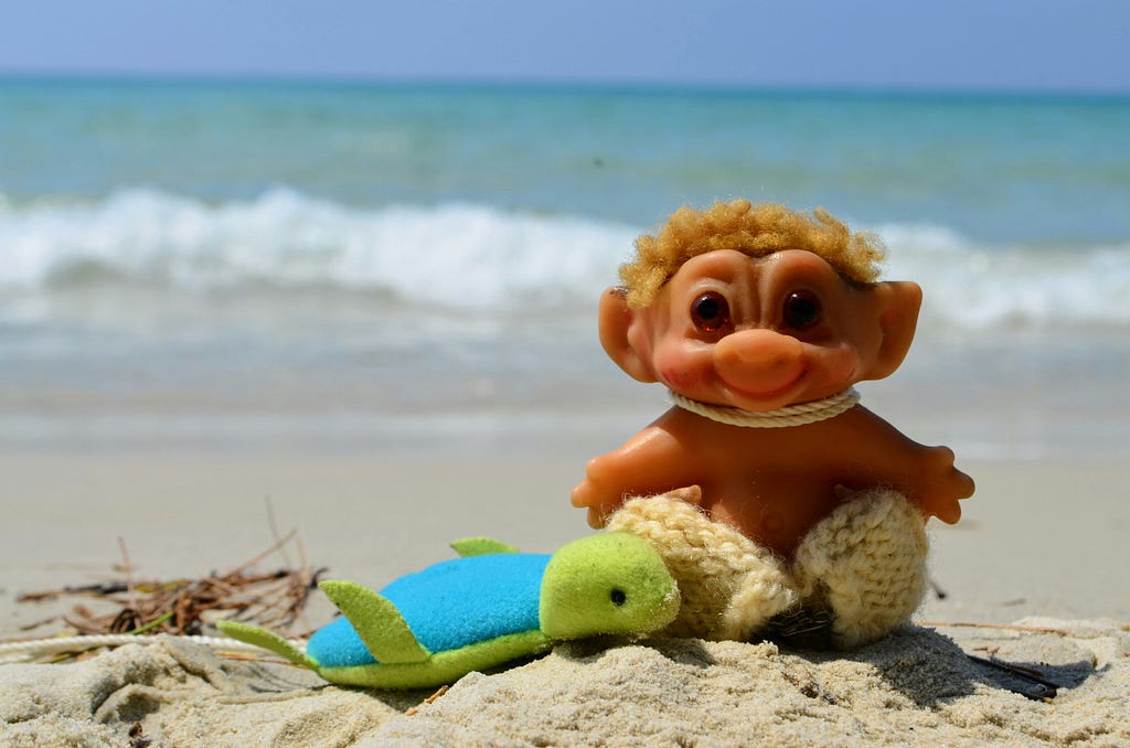 A cute troll children’s toy, sitting on the sand next to a toy surfboard, with the ocean in the background.