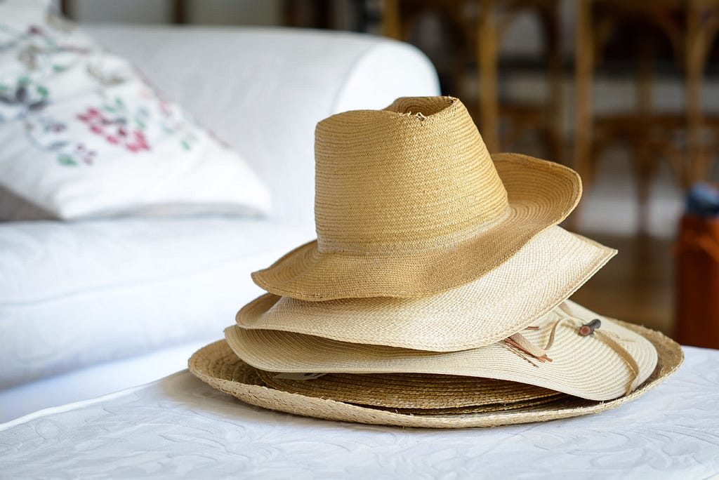 pile of straw hats on a perfect white ottoman. perfect white couch with mildly floral embroidered pillow is in the background.