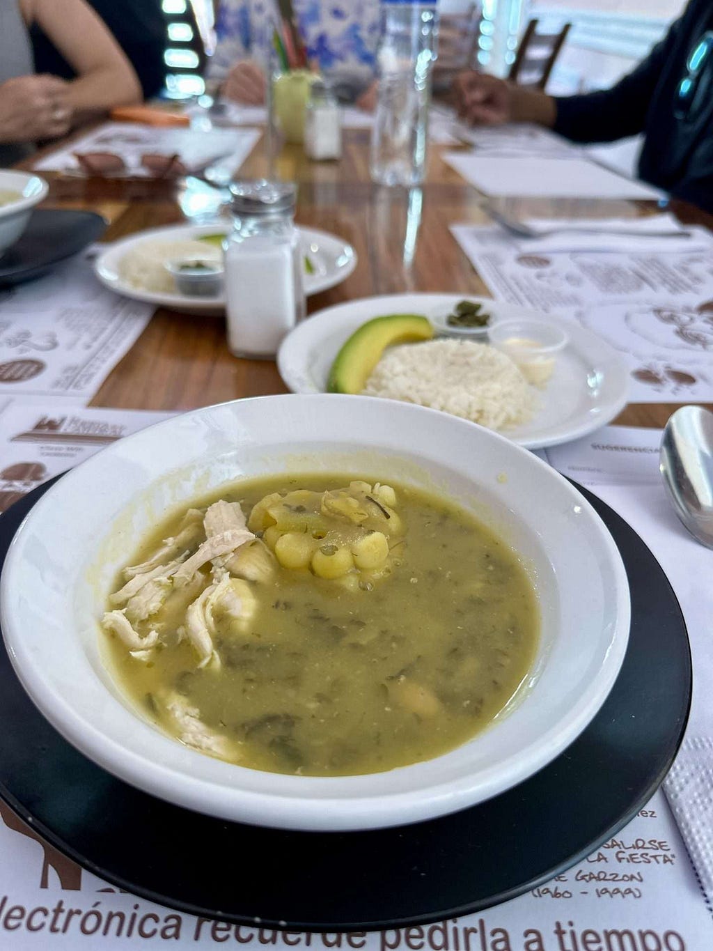 A bowl of ajiaco soup with corn on a table. Behind it, a plate with white rice, avocado slices, and a side dish of capers. Three people are seated at the table in the background.