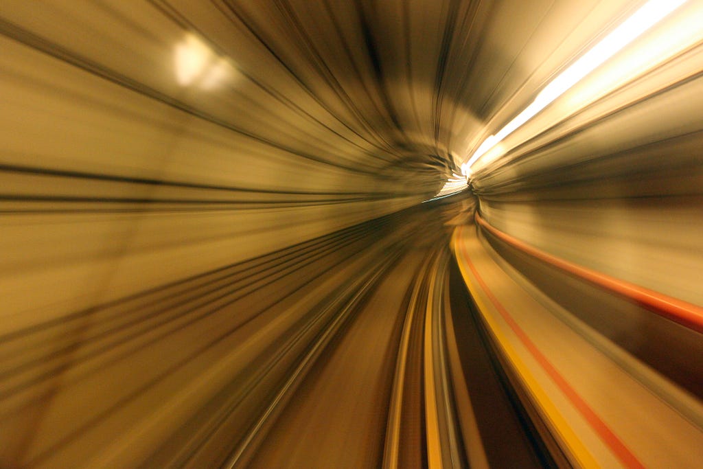 This image captures the dynamic visual effect known as “tunnel vision,” likely taken from the perspective of a moving vehicle inside a tunnel. The motion blur creates lines that recede rapidly towards a single point in the distance, simulating the high-speed movement through the tunnel. The walls and ceiling of the tunnel appear to stretch outwards due to the long exposure or slow shutter speed of the camera, intensifying the sense of speed and direction. Light sources within the tunnel create s