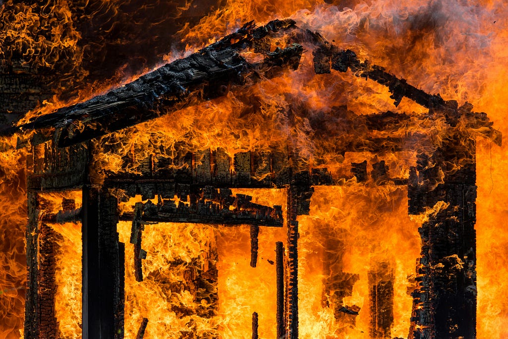 Image of a building on fire; it might have been a cabin. The only thing left and still ablaze is the framework, which appears blackened and ready to collapse. The flames are bright orange and seem out of control. Some dark grey smoke is also visible in the shot.