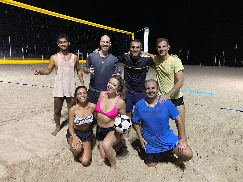 An image of smiling people on the beach at night in front of a volley net and holding a football