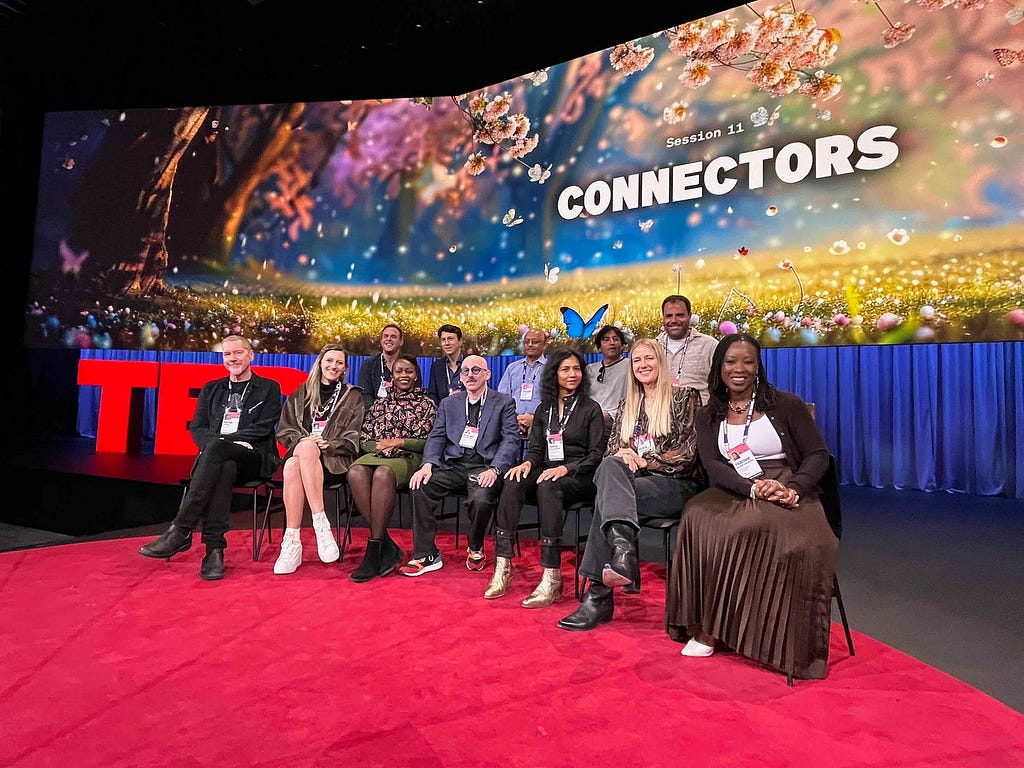 A photograph of the author with 11 other people seated together on the TED stage, in front of the TED letters and a screen that reads ‘Session 11: Connectors’