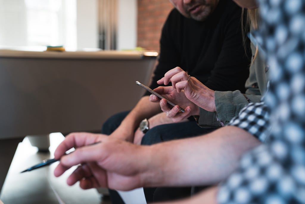 A close up view of a person using a mobile phone with 2 people looking at him.