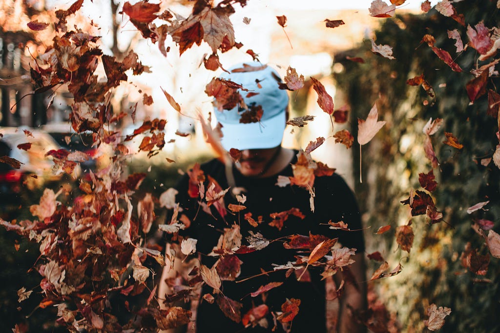 A man in a white baseball cap, pulled down low, is standing as fall leaves swirl around him.