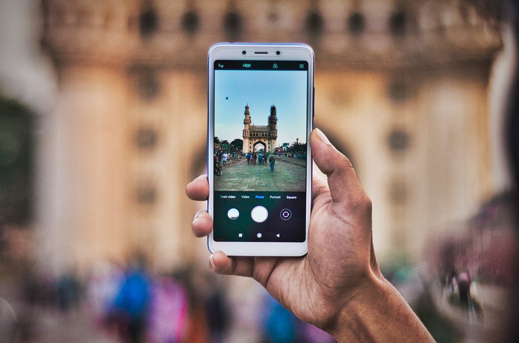 A person’s hand holds a phone and takes a picture of a famous landmark in India.