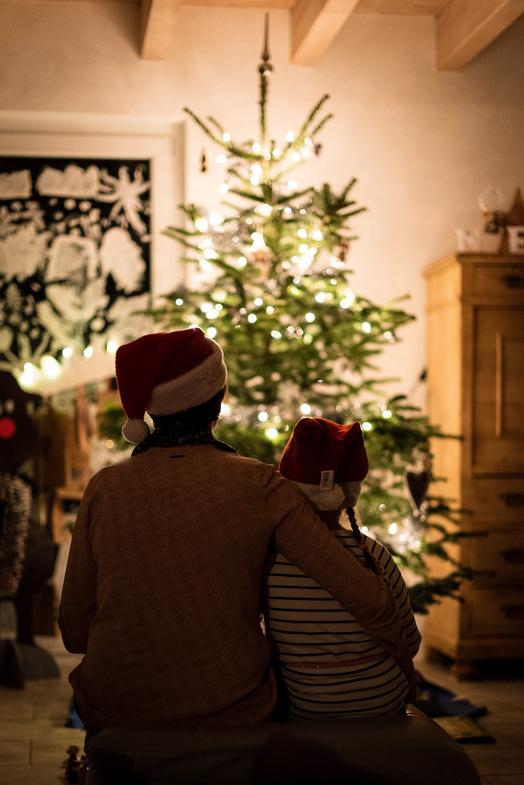 A parent and a child wearing Santa hats sitting in front of a small Christmas tree