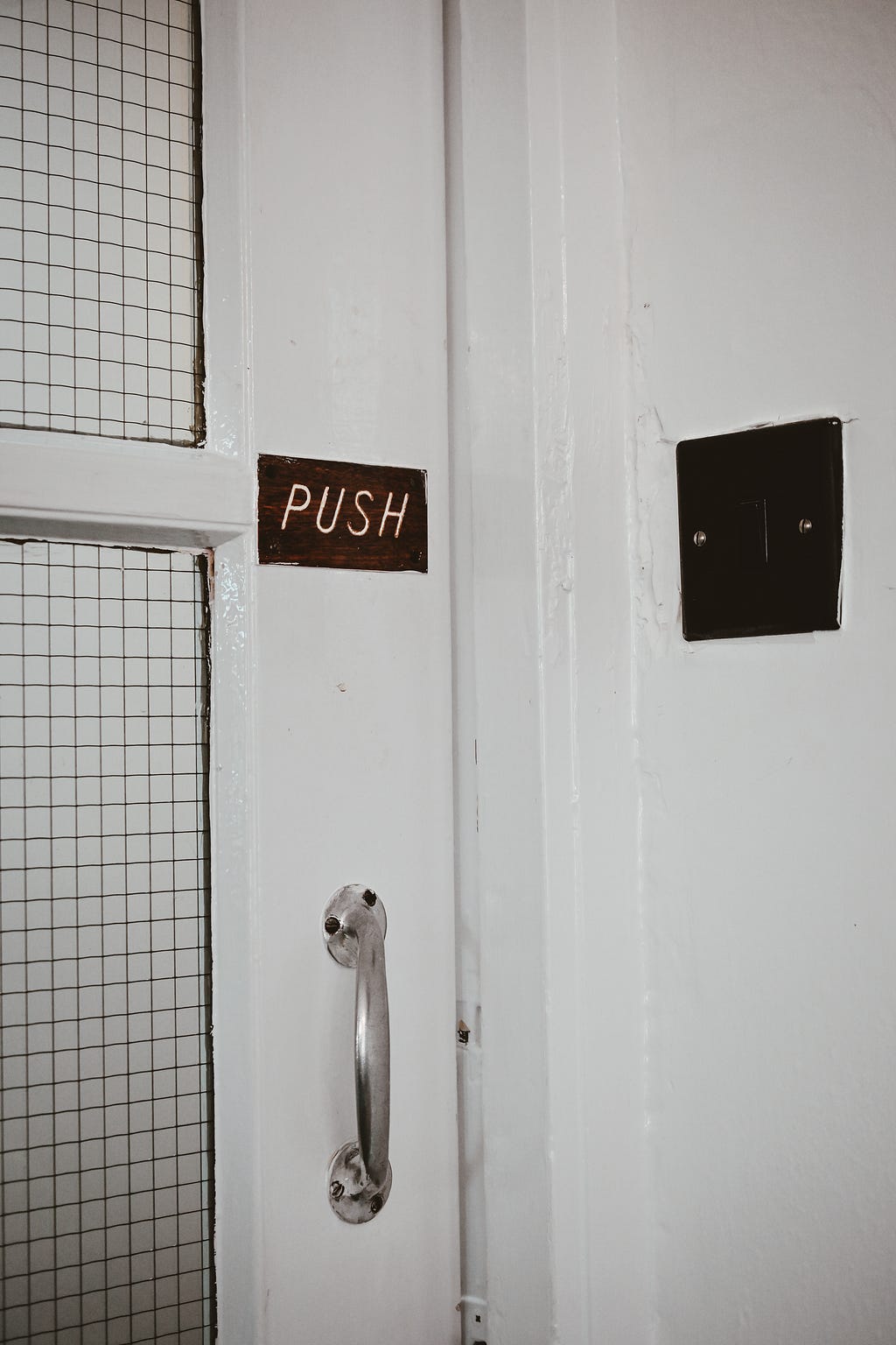 A white door with a curved handle facing outwards. Above the handle is a small sign that says, “Push.”