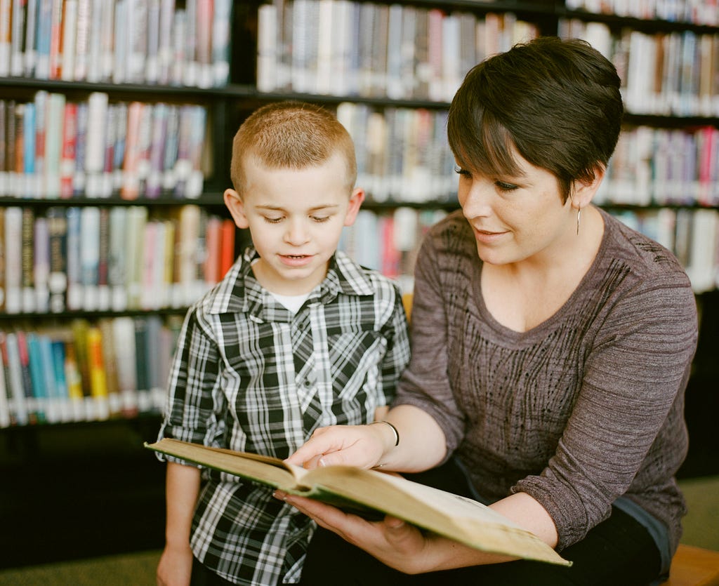 A woman is teaching a boy to read a text from a book in the library