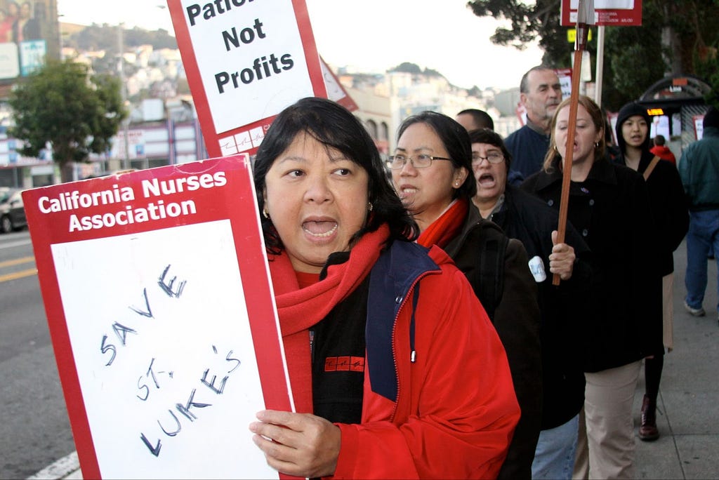 Filipina woman in red jacket and scarf standing on sidewalk holding a sign printed with California Nurses Association at the top and “Save St. Luke’s” handwritten in black pen in white space below. Other people are in background behind her holding signs.