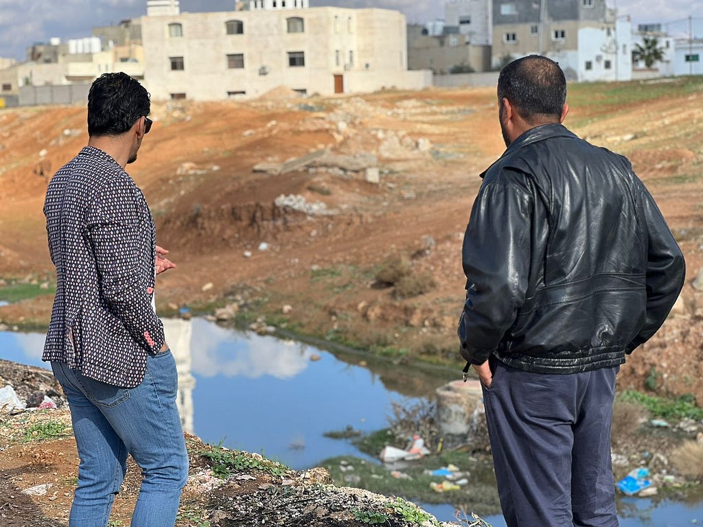 Two men with their backs to the camera look at a pool of water that is bordered by small piles of trash and is located downhill from a medium-sized building.