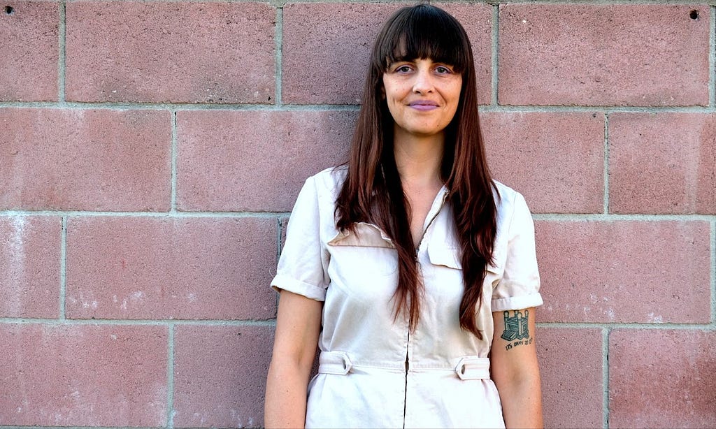 Sarah leans against a pinkish brick wall. They are light-skinned, femme non-binary, and wear a baby pink denim jumpsuit. They have long brown hair, bangs, and wear light purple lipstick. They are smiling, which makes dimples and soft lines around their mouth and eyes. The tattoo on their upper arm is of a bouncy castle, and beneath it in calculator font reads, “It’s okay to cry.”
