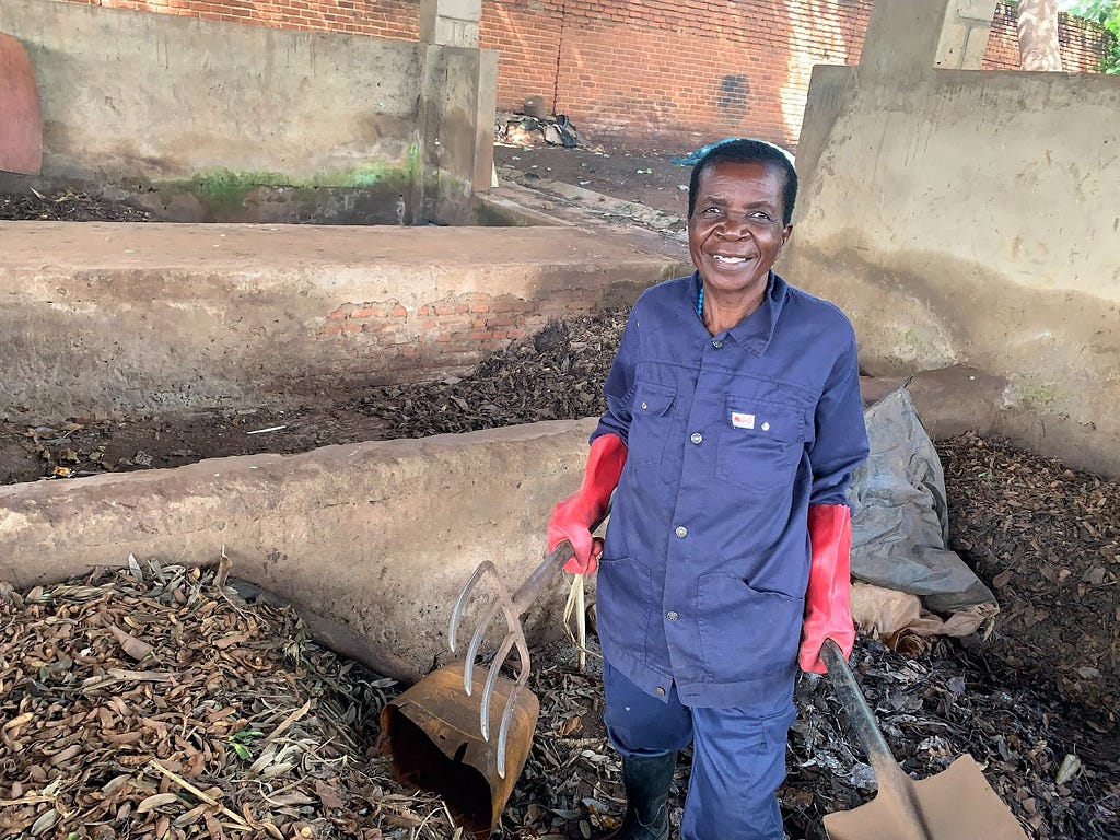 A woman wearing a blue uniform and red gloves up to her elbows, and holding a pitchfork and shovel, stands inside the manure-making site where she works.