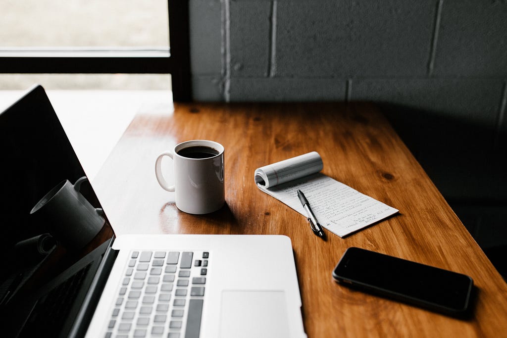 An image of a laptop sitting on a brown desk, a coffee mug, notepad with a pen and a phone.