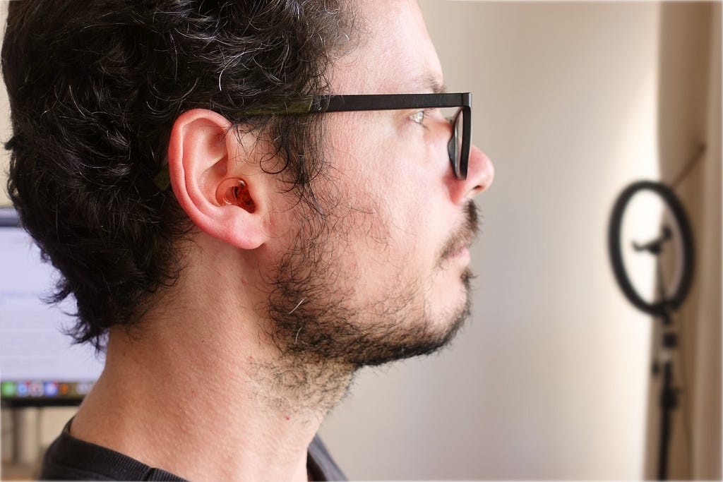 White man’s face, with glasses and beard, in profile, with emphasis on a pink plug in the ear.