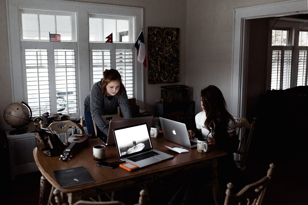 Two women are working on laptops at a kitchen table that is covered in mugs of coffee, a guitar, and other clutter.