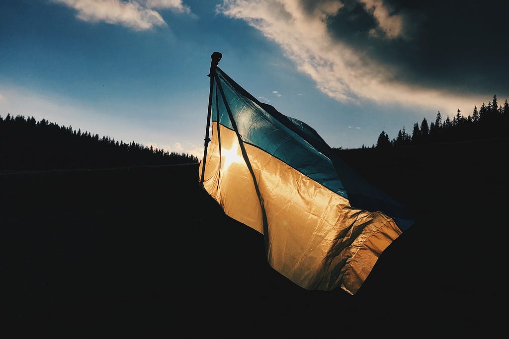 The Ukrainian flag with blue on top and yellow on bottom. Flag stands amidst background of clouds, sky, and trees in shadow.