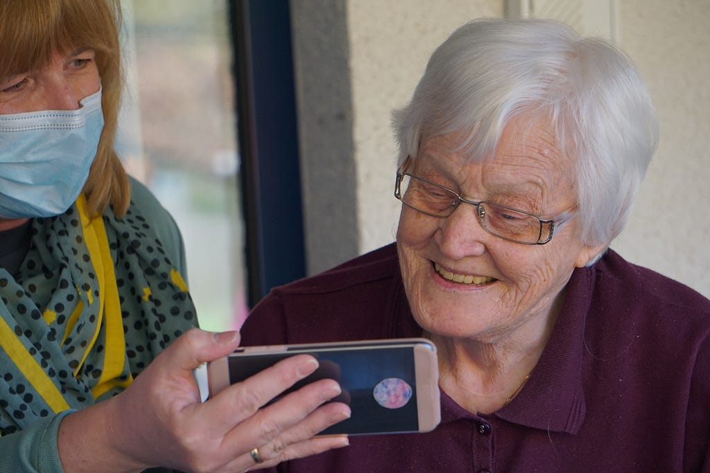 elderly woman smiles while looking at phone held by another woman wearing a mask
