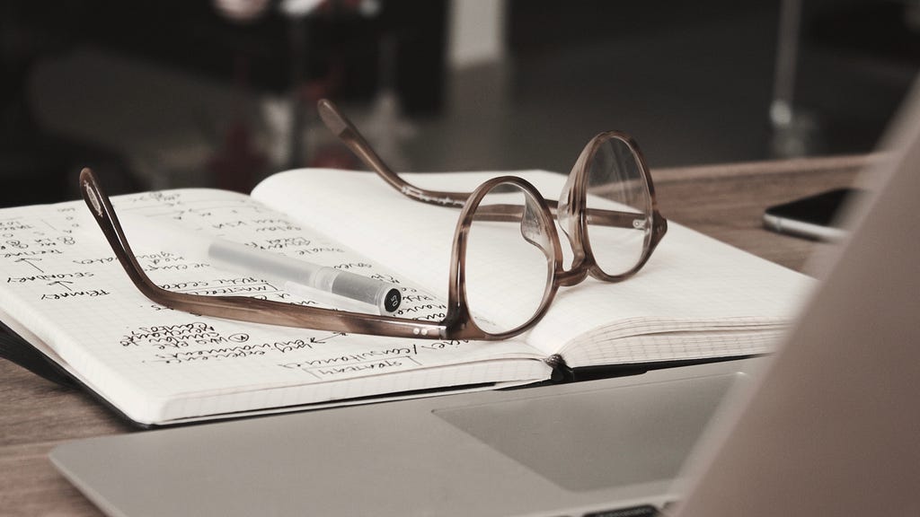 A pair of glasses and a pen on a notebook sitting in front of the laptop