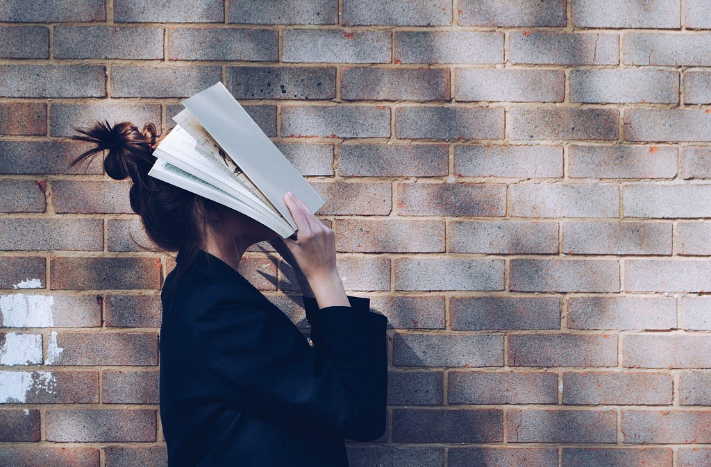 Uma mulher com um livro cobrindo o rosto sobre uma parede de tijolos.