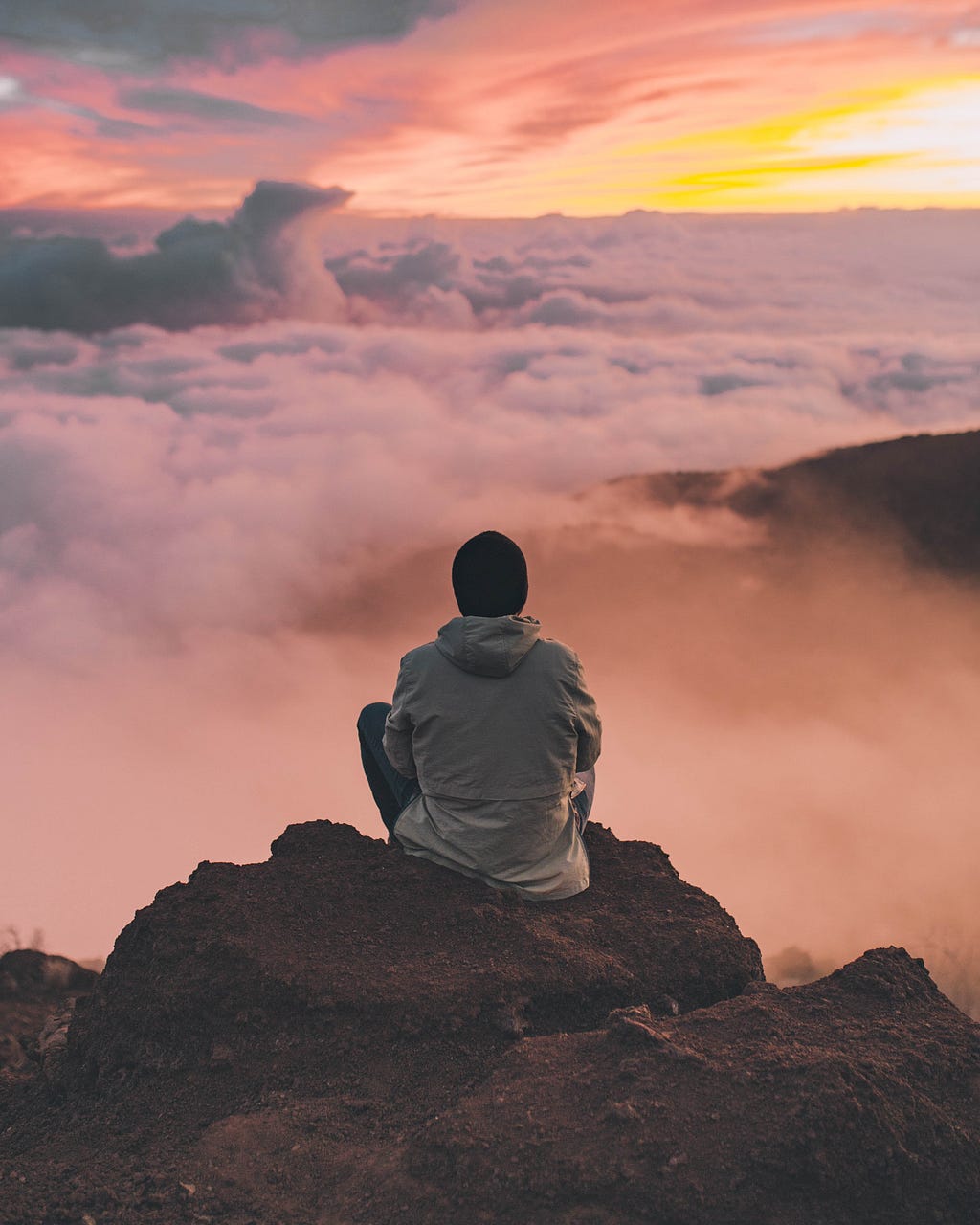 A man sitting on a mountain peak and staring at the clouds at sunset