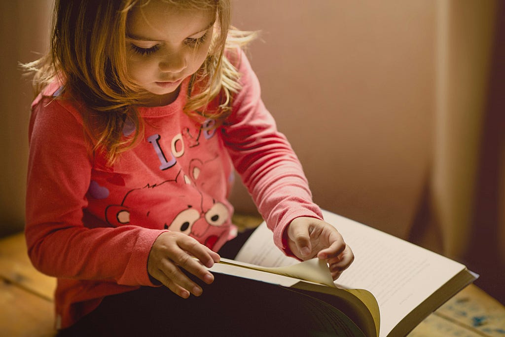 A young blonde girl wearing a pink shirt, printed with a cartoon face and the word Love, is seated on the floor turning pages in a large book on her lap.