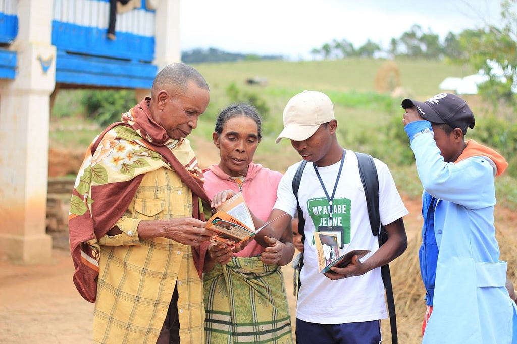 A man and woman who are parents to a teenager hold a magazine in their hands while a youth educator and his colleague from Projet Jeune Leader speak to them. The group is standing outside in what appears to be a rural or suburban setting of Madagascar.