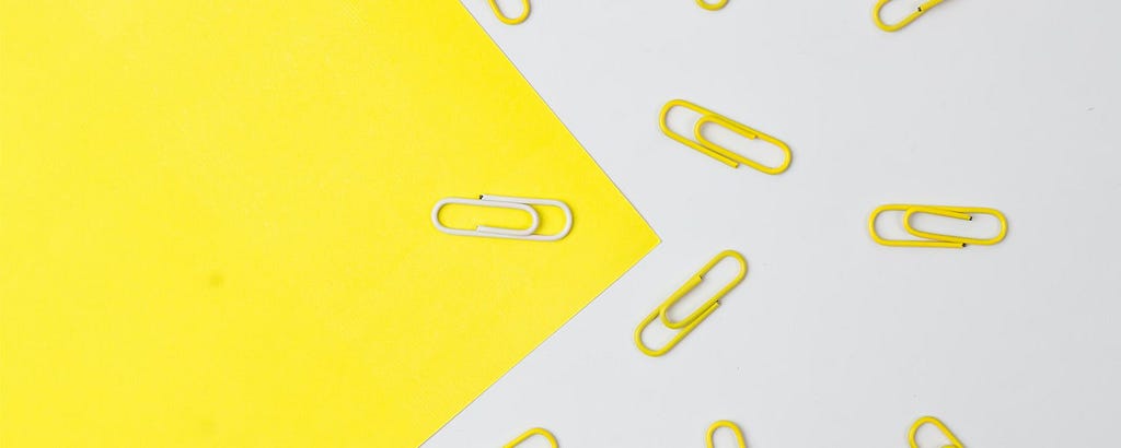 Piece of yellow paper on an angle with one white paperclip on it against a white background with many yellow paperclips on it