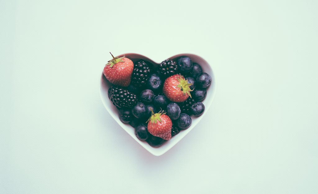 strawberries, blackberries and blueberries in heart-shaped bowl on green background