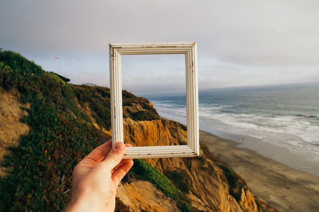 A hand holding empty frame up to a view of the ocean and mountains.