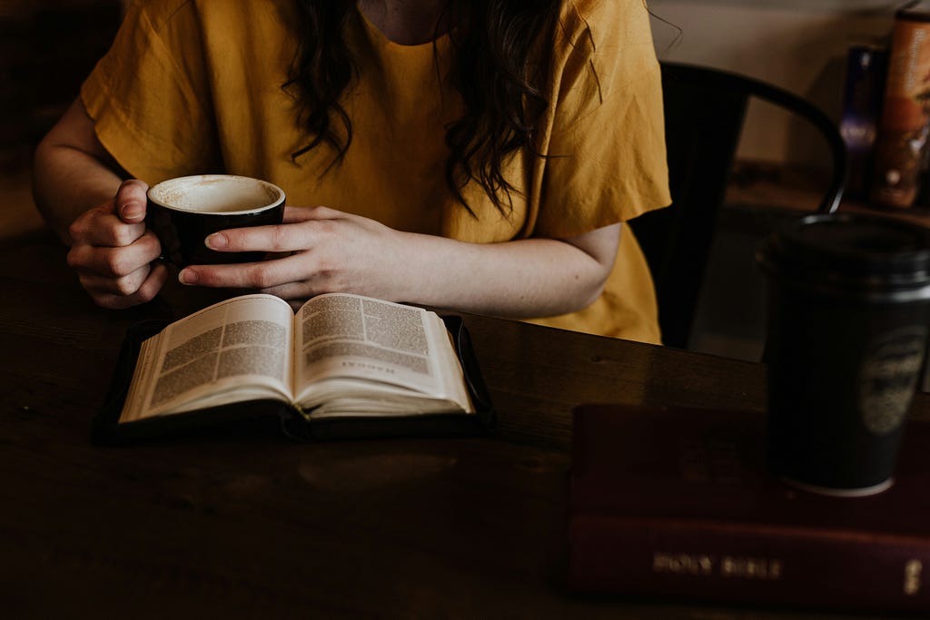A girl sits alone in a chair, reading a book while holding a cup of coffee.