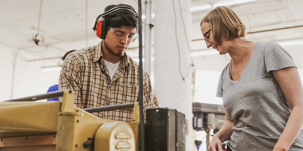 A woodworking instructor looks on as her student works on a project.