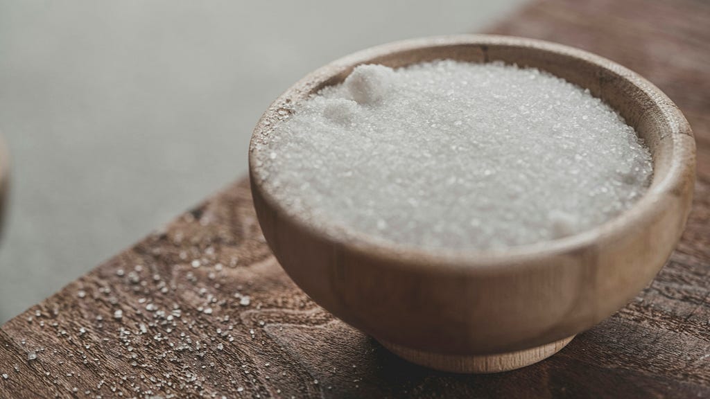White sugar in a small pale wooden bowl, on a grainy wood table top.