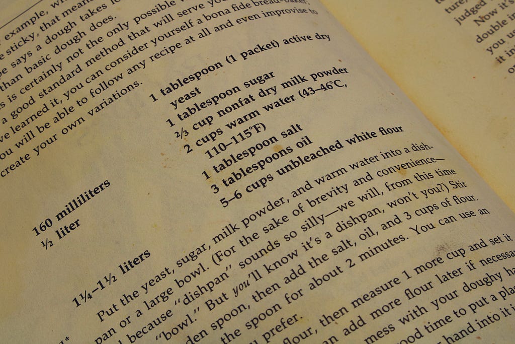 A page in a cookbook, with most of the page left unseen. What’s visible shows an ingredient lists and recipe text for what appears to be dough. The book is flat and open.