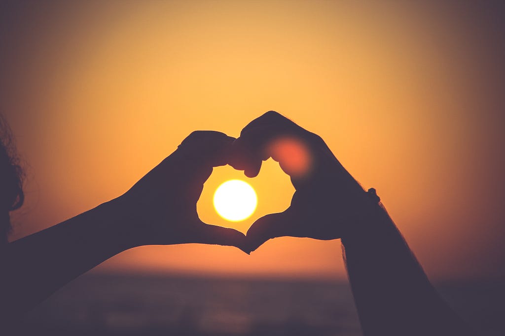 Two people joining hands to make a heart shape, while the sun is setting, with the setting sun in the center of that heart.