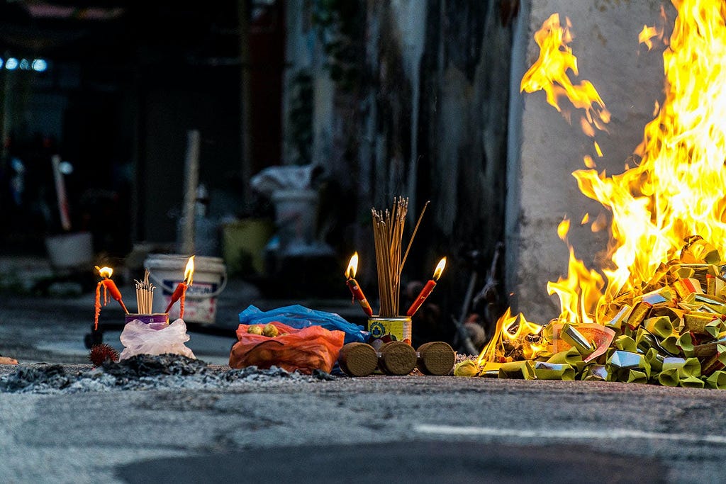 A fiery blaze consumes paper offerings during the Hungry Ghost Festival, with lit incense sticks and candles in front and a pile of vibrant paper effigies waiting to be offered against an early evening backdrop.