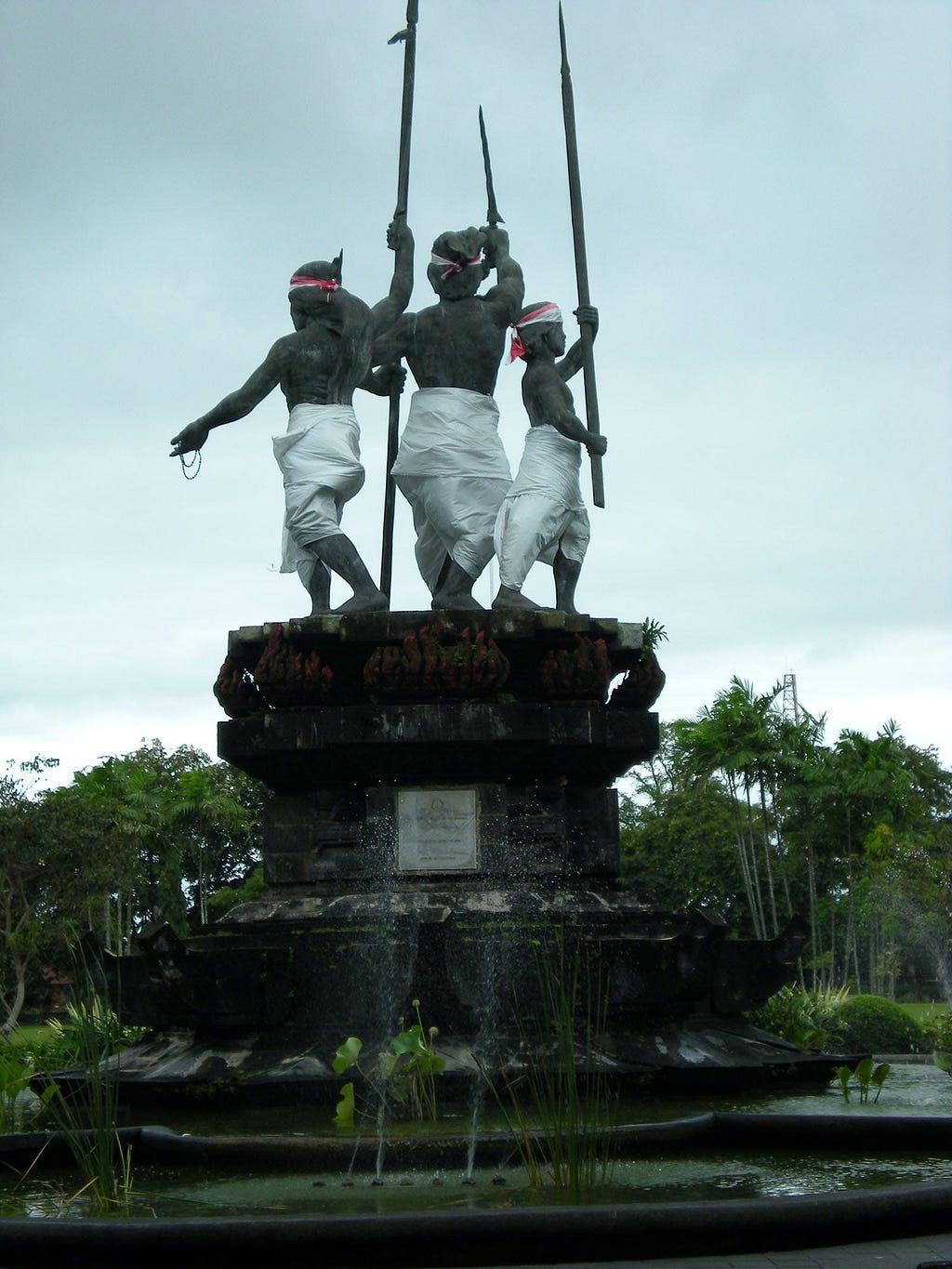 Monument in Denpasar, Bali, commemorating the 1906 Puputan, depicting three Balinese warriors in traditional attire, with raised spears, standing back-to-back on a pedestal against a cloudy sky. The monument is surrounded by a water feature with spouts and greenery.