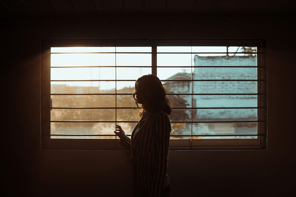 A woman standing in a dark room staring outisde through the blinds of a window.