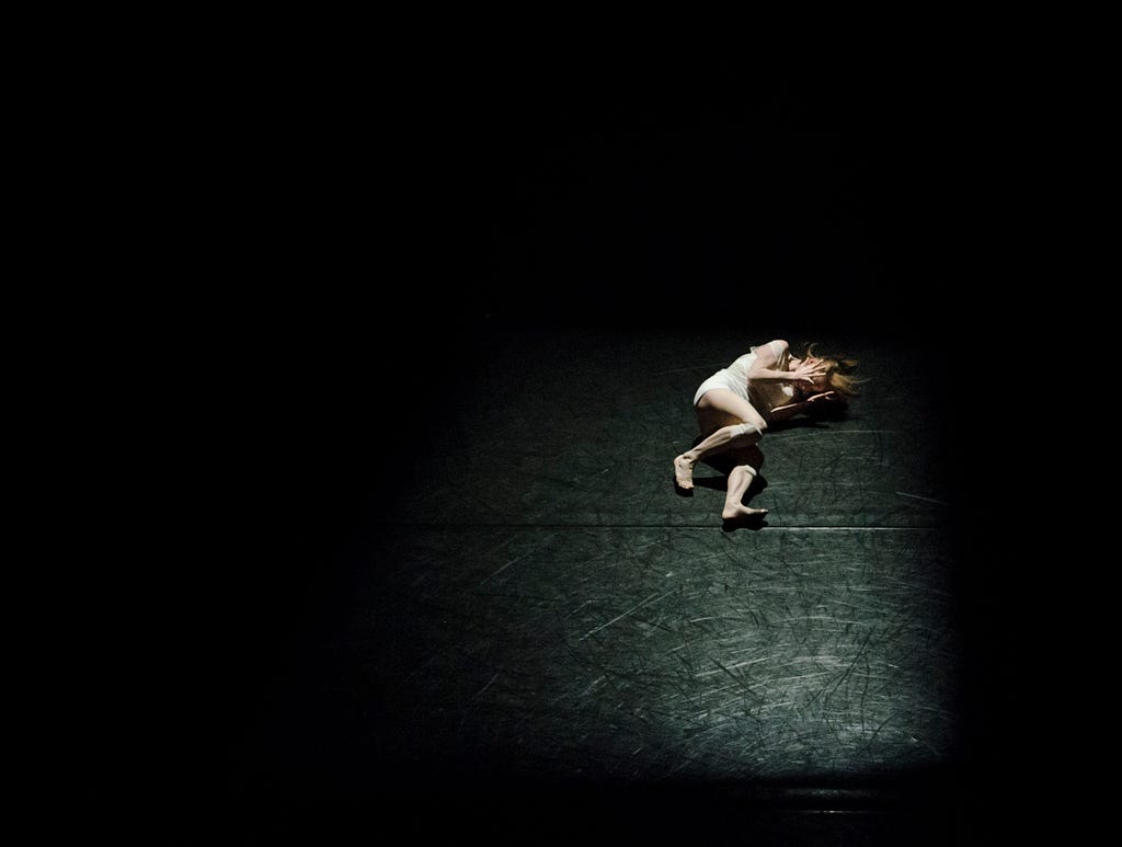 Image of a woman laying on her side on the dark ground. The image is black, with only a portion visible. The woman’s hands are near her face, and her legs are bent forwards, the top knee partly bent. She’s alone, and appears to be in pain.