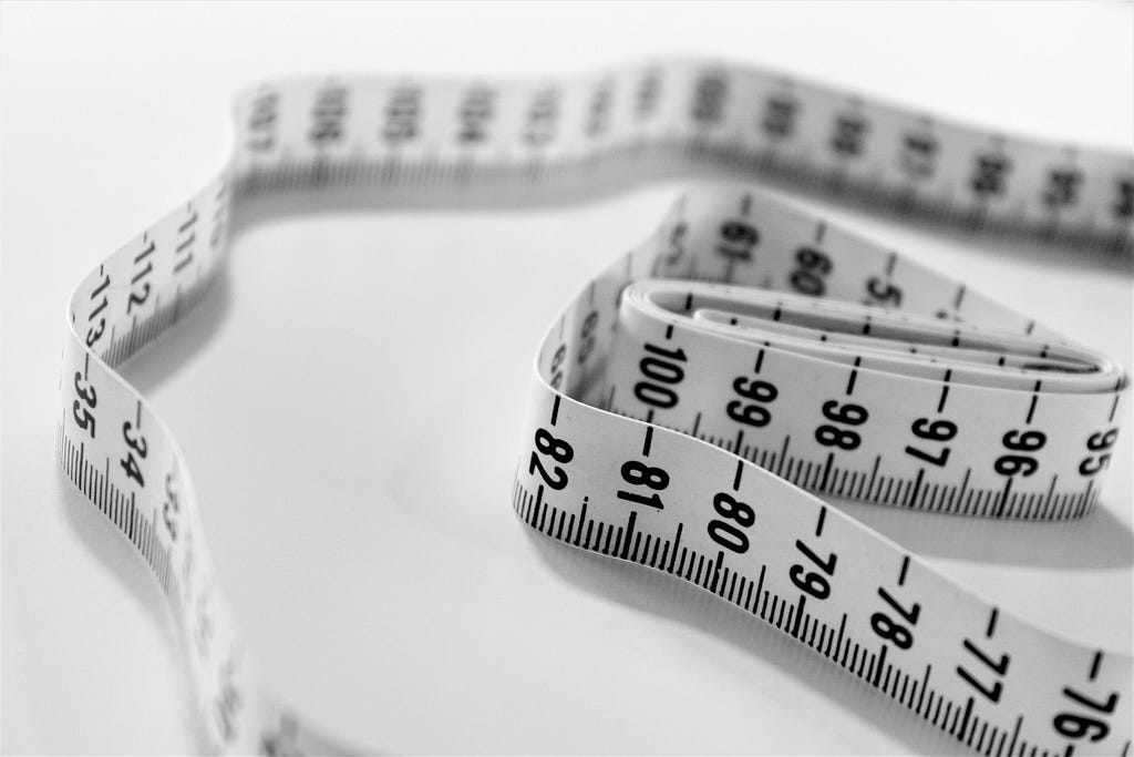 A close-up, black and white image of a measuring tape loosely coiled on a white background, with the numbers on the tape clearly visible and in focus