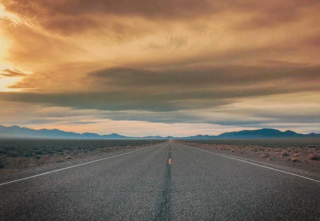A photo of an empty, open road in the middle of nowhere. The sky is cloudy with colors ranging from orange to blue. Mountains far in the distance.