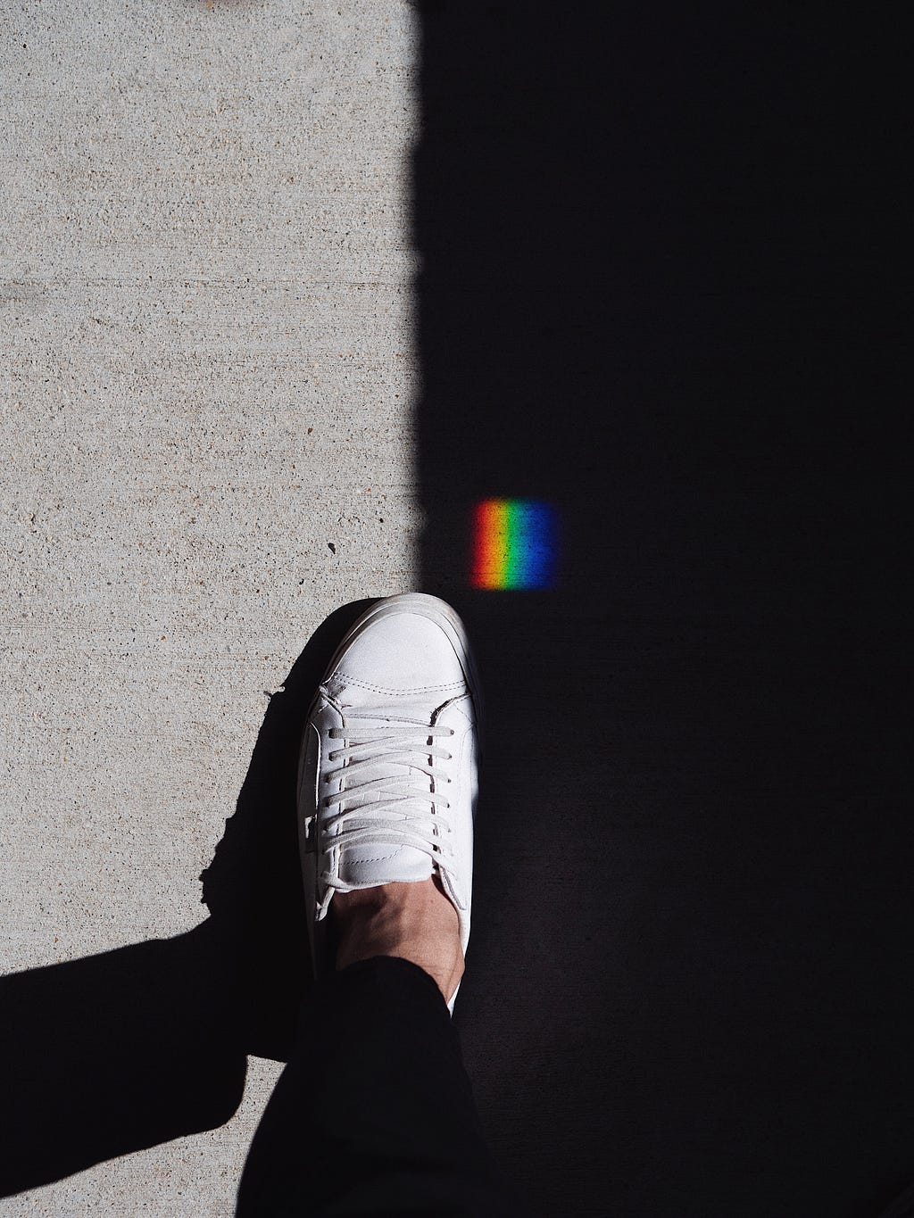 A white shoe on a pavement bisected by a dark, black shadow. To the left is light-gray pavement. To the right is the shadow. Above the show is a small square patch of rainbow light.