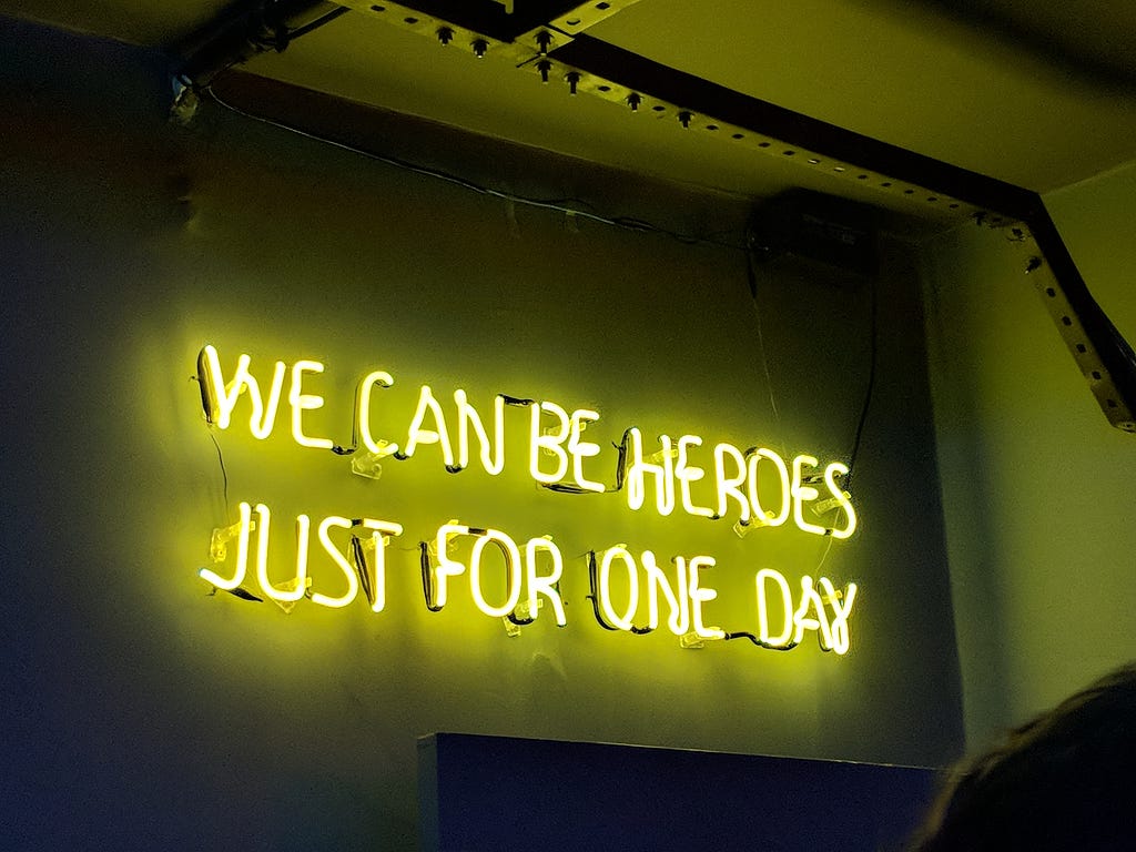 Neon sign on a wall in a dark room. The sign says ‘We can be heroes just for one day’