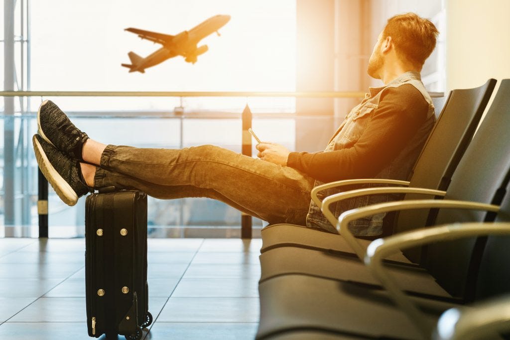 Man sitting in airport terminal with feet up on suitcase looking out the window as a plane takes off.