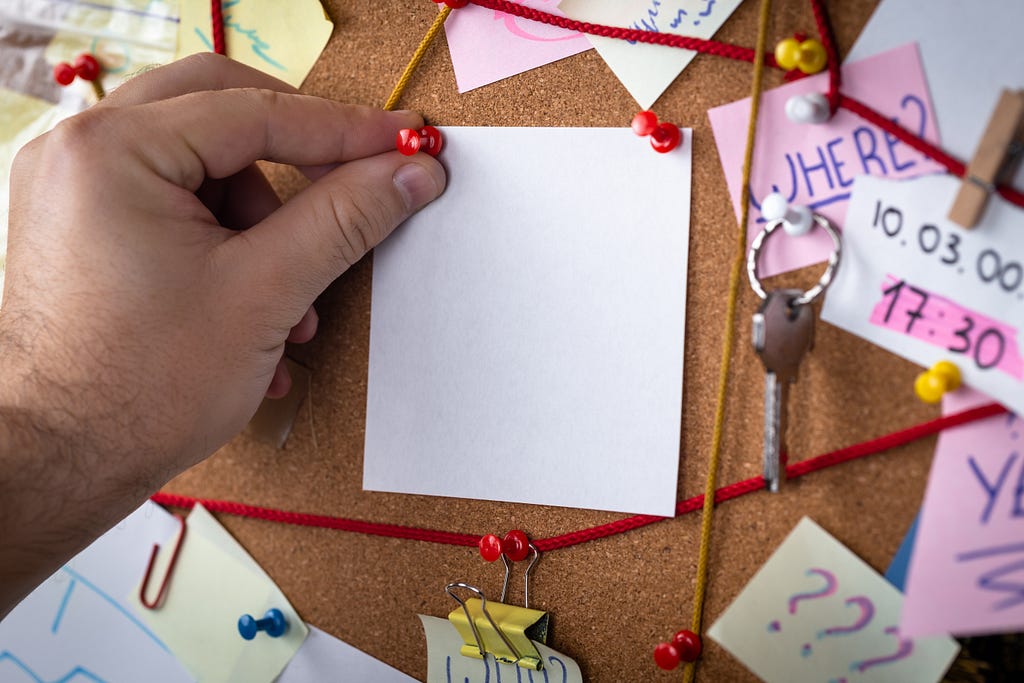 A hand pins a note into full cork noticeboard. Some notes are connected together with red string.
