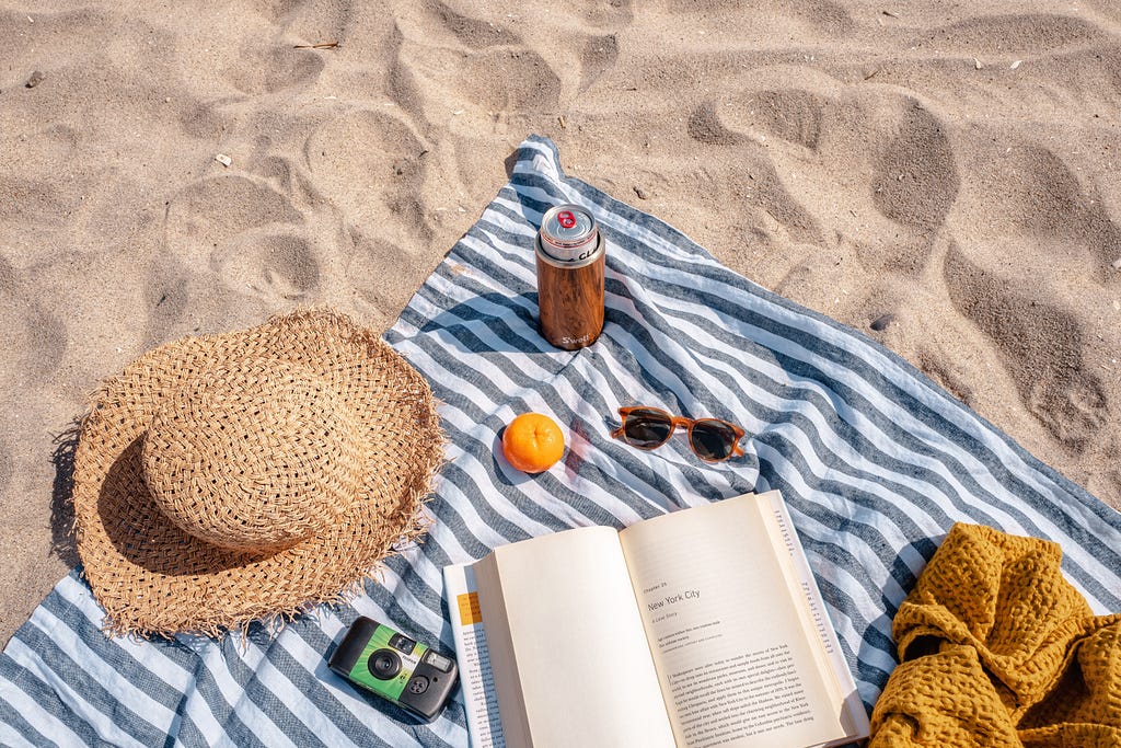 A beach towel on a sandy beach with book, a beer , shades and a camera.