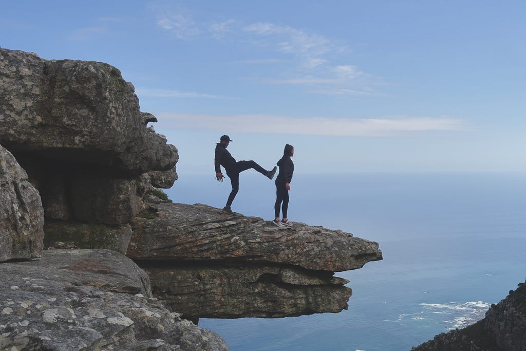 A man has his leg extended, preparing to kick a woman off a rock into the water