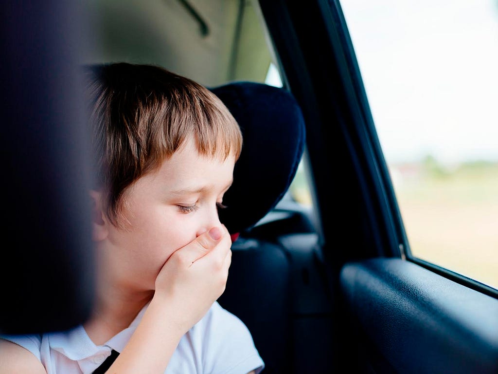 A child sitting in a car facing the window and covering his mouth. He looks ot have carsickness.