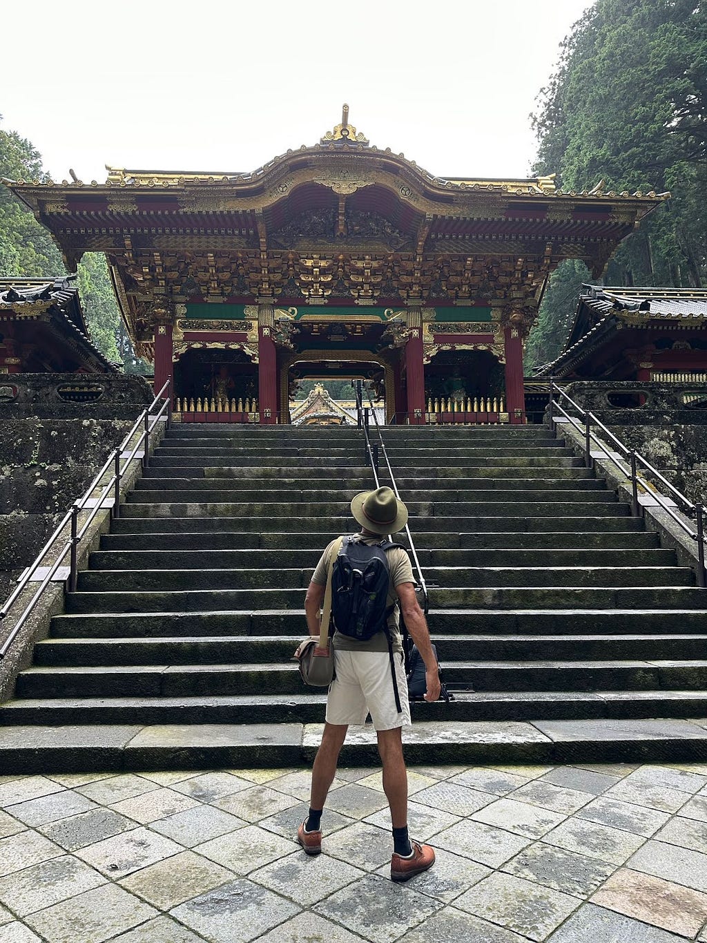 Man photographed from behind in green t-shirt, white shorts and with a small black backpack and green side bag. Green felt fedora. Looking off at the steps leading to a Japanese Buddhist temple.